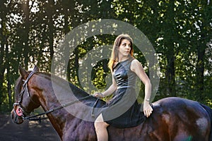 Young slim woman brunette in a black dress sitting on dark brown horse.