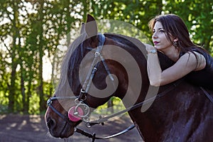 Young slim woman brunette in a black dress sitting on dark brown horse. Sunny summer evening