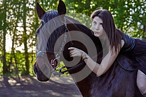 Young slim woman brunette in a black dress sitting on dark brown horse. Sunny summer evening.