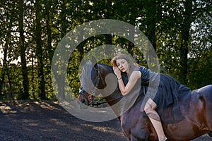 Young slim woman brunette in a black dress sitting on dark brown horse. Sunny summer evening.