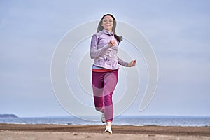 Young slim woman brunette in sportswear runs along the sandy bank of a large river.