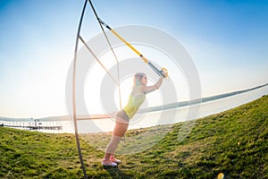 Young slim  woman doing suspension training with fitness straps outdoors near the lake at daytime. Healthy lifestyle