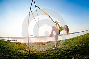 Young slim  woman doing suspension training with fitness straps outdoors near the lake at daytime. Healthy lifestyle