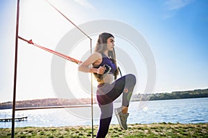 Young slim  woman doing suspension training with fitness straps outdoors near the lake at daytime. Healthy lifestyle