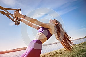 Young slim  woman doing suspension training with fitness straps outdoors near the lake at daytime. Healthy lifestyle