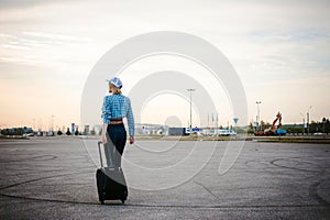 Young slim woman dressed in blue checked shirt, cap and jeans, Will depart at dawn on summer day on freedom trip with black travel