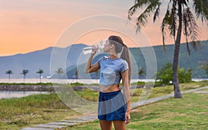 Young slim woman drinking water after exercises and sport against sunset and beach with palms