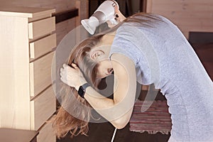 Young slim woman in gray dress dries her light brown hair with hair dryer, head down. Morning routine, interior in beige color, wo