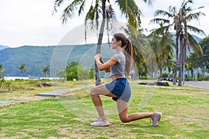 Young slim woman squatting and doing sport exercises against sunset and beach with palms