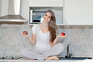 Young slim woman standing in kitchen on table, holding red apples, looking