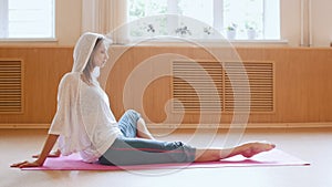 Young slim woman in white hoodie sitting on the yoga mat and pulling her toes forward