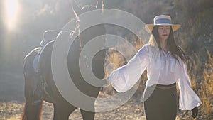 Young slim woman in white shirt and straw hat walking in slow motion holding horse bridle. Portrait of confident