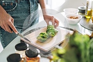 Young slim woman in white t-shirt and blue jeans cooking healthy food with cucumber in kitchen at home