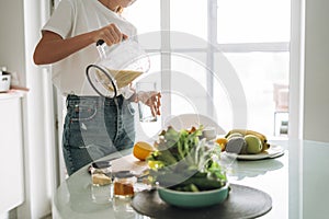 Young slim woman in white t-shirt and blue jeans pouring fruit smoothie healthy food in kitchen at home