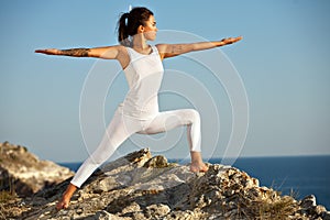 Young slim Yoga woman in zen meditating in warrior pose relaxing outside on top of mountains and sea on sunrise or sunset.