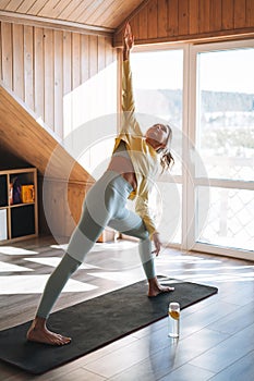 Young sllim fitness blonde woman practice morning yoga near window of home