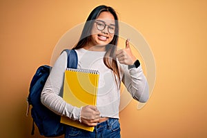 Young smart asian student girl wearing backpack holding notebook over yellow background happy with big smile doing ok sign, thumb
