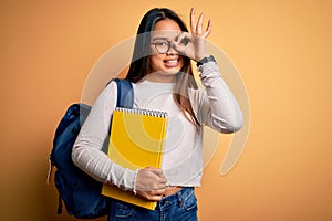 Young smart asian student girl wearing backpack holding notebook over yellow background with happy face smiling doing ok sign with