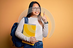 Young smart asian student girl wearing backpack holding notebook over yellow background surprised with an idea or question