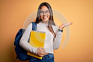 Young smart asian student girl wearing backpack holding notebook over yellow background very happy and excited, winner expression