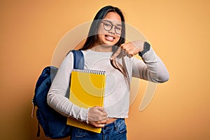 Young smart asian student girl wearing backpack holding notebook over yellow background very happy pointing with hand and finger