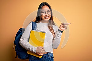 Young smart asian student girl wearing backpack holding notebook over yellow background very happy pointing with hand and finger