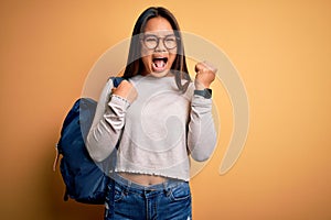 Young smart asian student girl wearing backpack standing over isolated yellow background screaming proud and celebrating victory