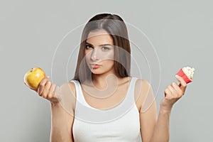 Young smart woman choosing between healthy fruits and tasty cakes on grey background