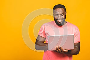 Young smiling african american man standing and using laptop computer isolated over yellow background