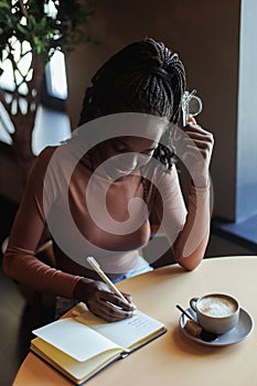 Young smiling African-American woman sitting at table with cup of coffee cappuccino in cafe, writing text in notebook.