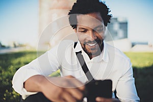 Young smiling African man sending text message from smartphone while sitting at sunny city park.Concept of happy