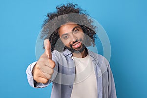 Young smiling Arabian man shows thumbs up and looking at camera stands in studio