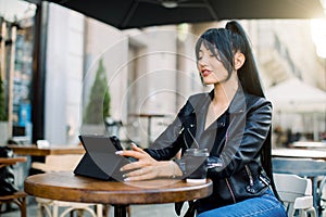 Young smiling Asian brunette girl with ponytail hair, using digital tablet for work while sitting at outdoor cafe