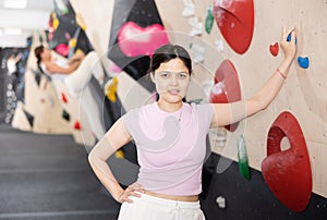 Young smiling Asian girl coach stands at climbing wall and leans holding on to hook.