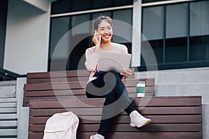 Young smiling Asian girl using a mobile phone and laptop on her lap in university campus building