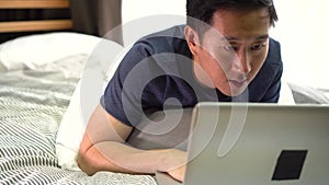 Young smiling Asian man in casual clothing and lying on bed with laptop computer. Male freelancer working from home on