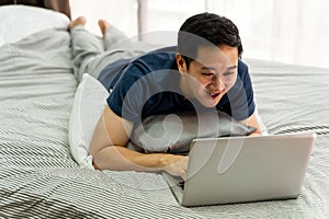 Young smiling Asian man in casual clothing and lying on bed with laptop computer. Male freelancer working from home on