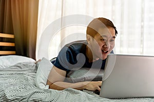 Young smiling Asian man in casual clothing and lying on bed with laptop computer. Male freelancer working from home on