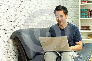 Young smiling Asian man in casual clothing on sofa working on laptop computer. Male freelancer working from home on