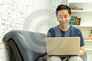 Young smiling Asian man in casual clothing on sofa working on laptop computer. Male freelancer working from home on