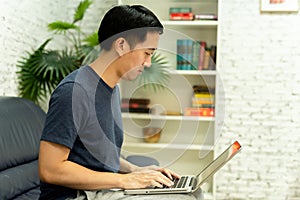Young smiling Asian man in casual clothing on sofa working on laptop computer. Male freelancer working from home on