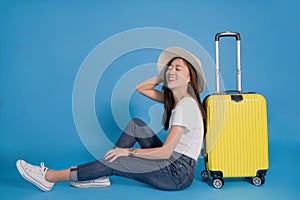 Young smiling Asian woman traveler is happy sitting near yellow suitcase on blue background. She used her hand to hold her hat