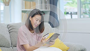 Young smiling Asian woman using tablet buying online shopping by credit card while lying on sofa when relax in living room at home