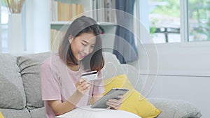 Young smiling Asian woman using tablet buying online shopping by credit card while lying on sofa when relax in living room at home