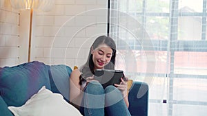 Young smiling Asian woman using tablet buying online shopping by credit card while lying on sofa when relax in living room at home