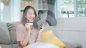 Young smiling Asian woman using tablet buying online shopping by credit card while lying on sofa when relax in living room at home
