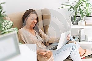 Young smiling attractive woman in cozy beige cardigan working at laptop sitting in chair at the home with green house plants