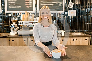 Young smiling barista girl with cup of freshly prepared coffee. Blond woman in an apron, near the bar counter in coffee shop