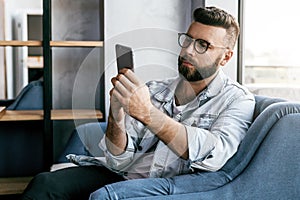 Young smiling bearded businessman is sitting in cafe, using smartphone. Freelancer work in coffee shop.Online education