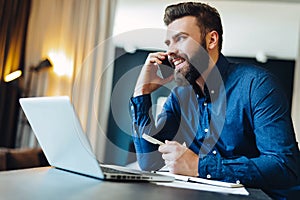 Young smiling bearded businessman sitting in front of computer, talking on cell phone, holding pen. Phone conversations. photo